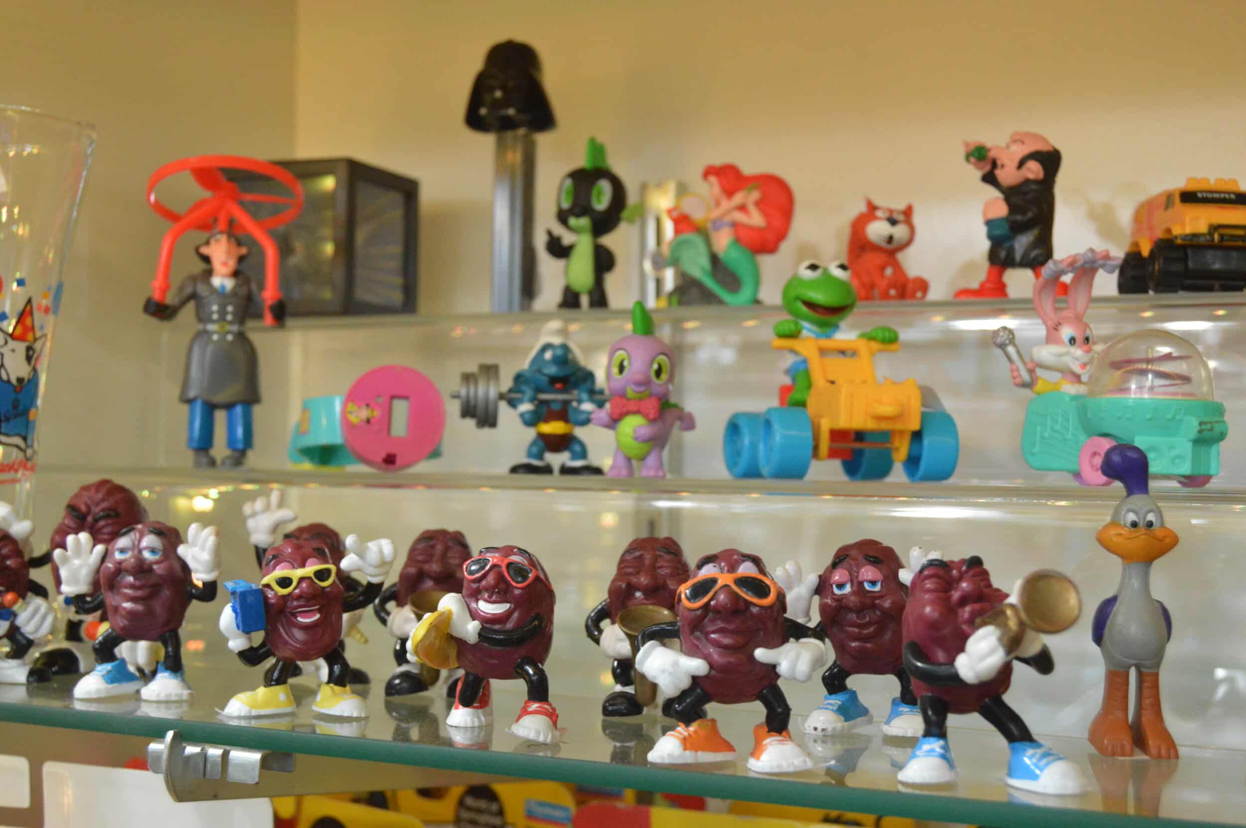 A row of dancing California Raisins figurines, in various dancing postures, some of them wearing sunglasses or playing musical instruments, occupies a shelf at Meep Meepleton’s. More figurines are arrayed on two shelves above the raisins, including an Inspector Gadget and a figure of Kermit the Frog riding on a wheeled vehicle.