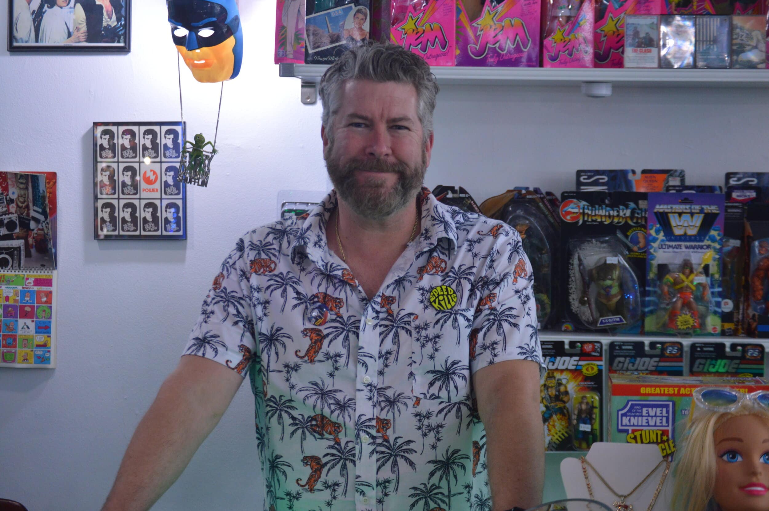 Dave Farrar, co-owner of Meep Meepleton’s World of Fun, stands in his shop wearing a shirt printed with palm trees.