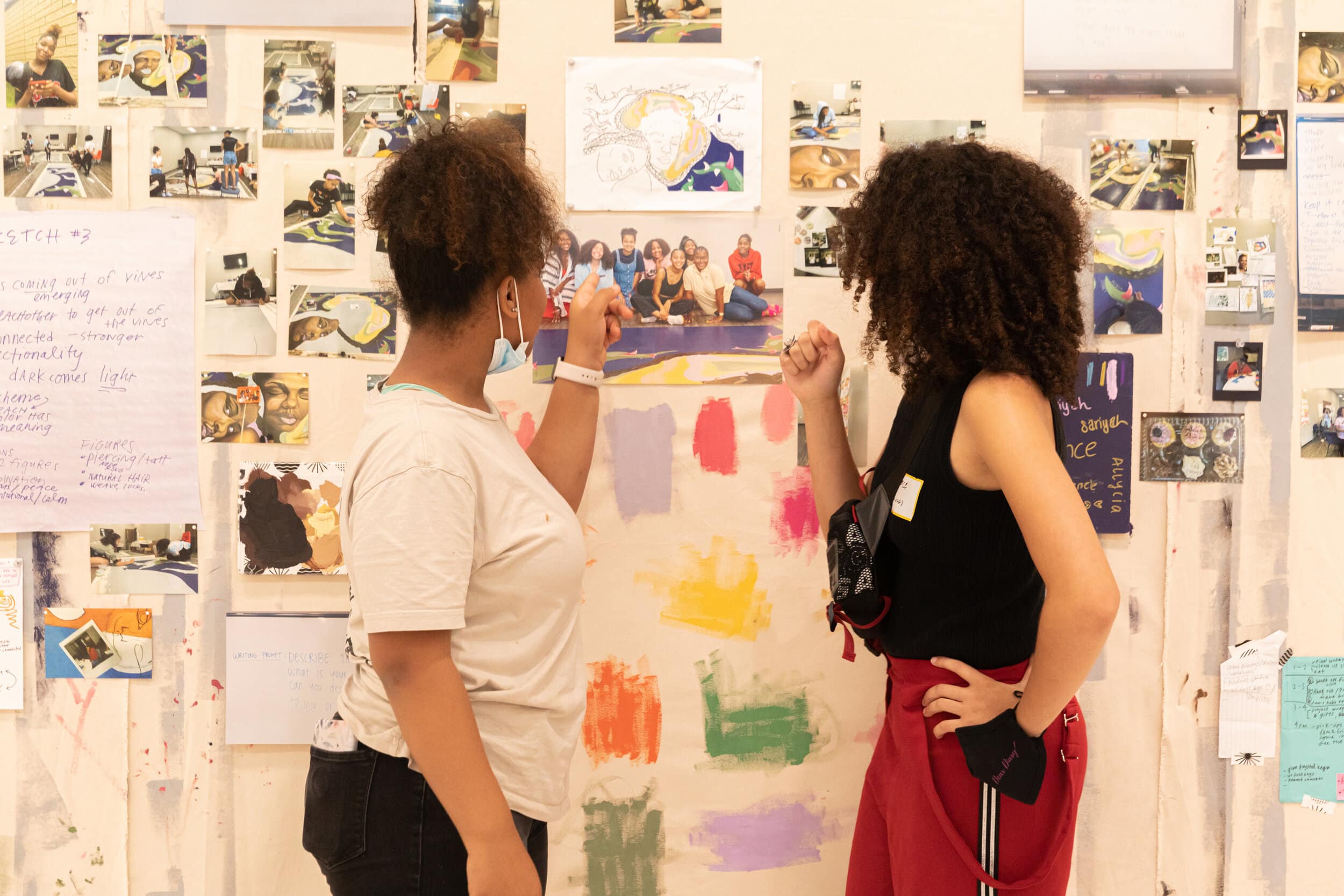 Kadence and Aviana, two of the Dear Diary teen artists, stand in front of the artmaking process wall looking at a photograph of the artists group. Photographs, notes, color samples, and other images are pinned to the wall. Photo by Amadou Kromah.