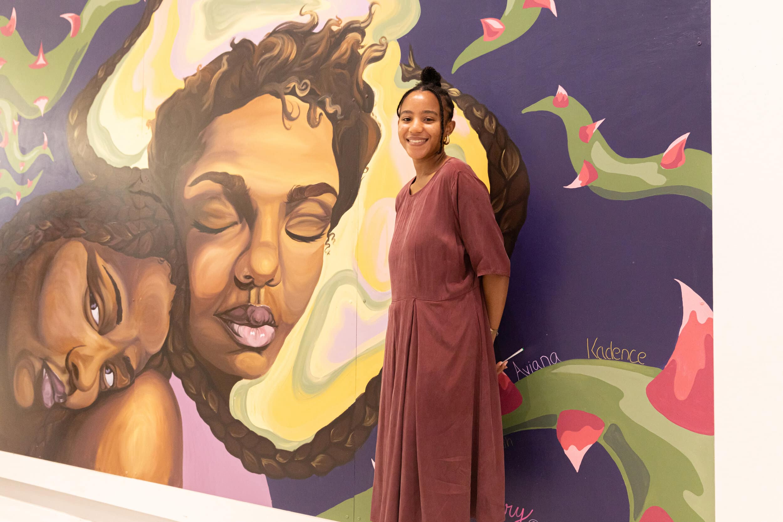 Artist Shiloah Symone Coley stands in front of the Crowns In Chaos mural, smiling. Photo by Amadou Kromah.