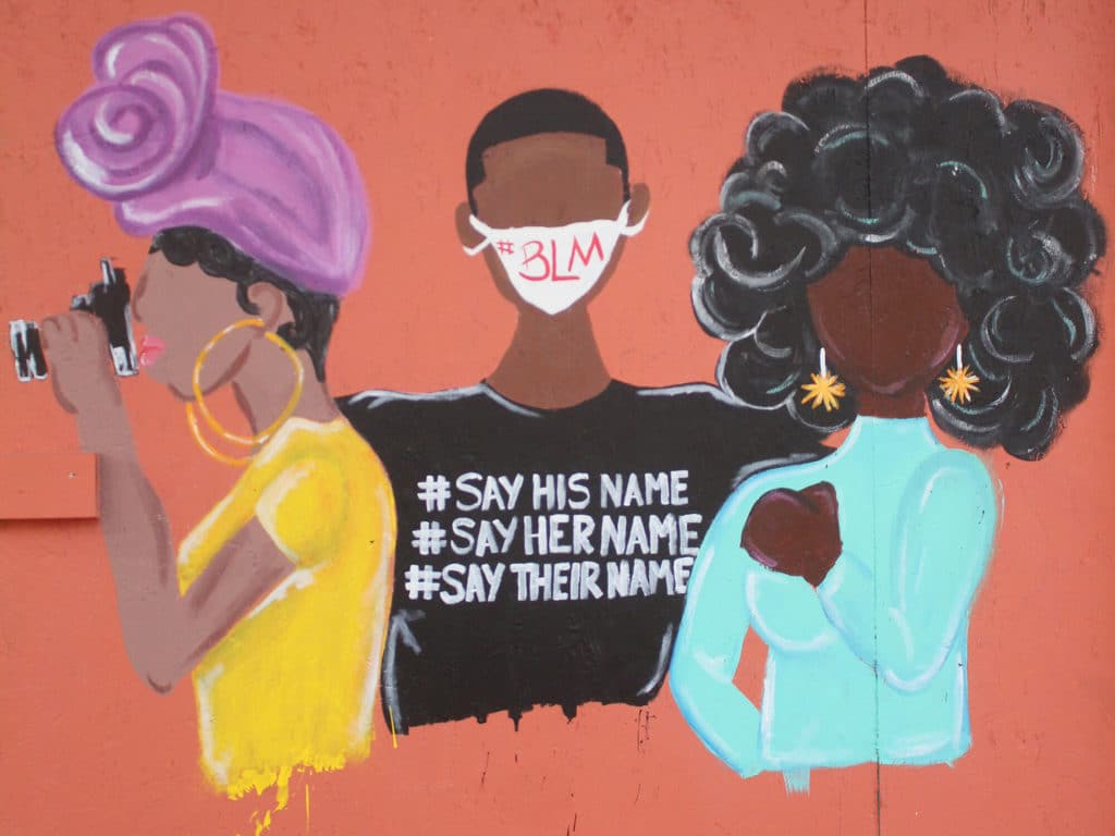 A photo shows a street mural depicting three Black women. The figure in the middle is wearing a face masks with