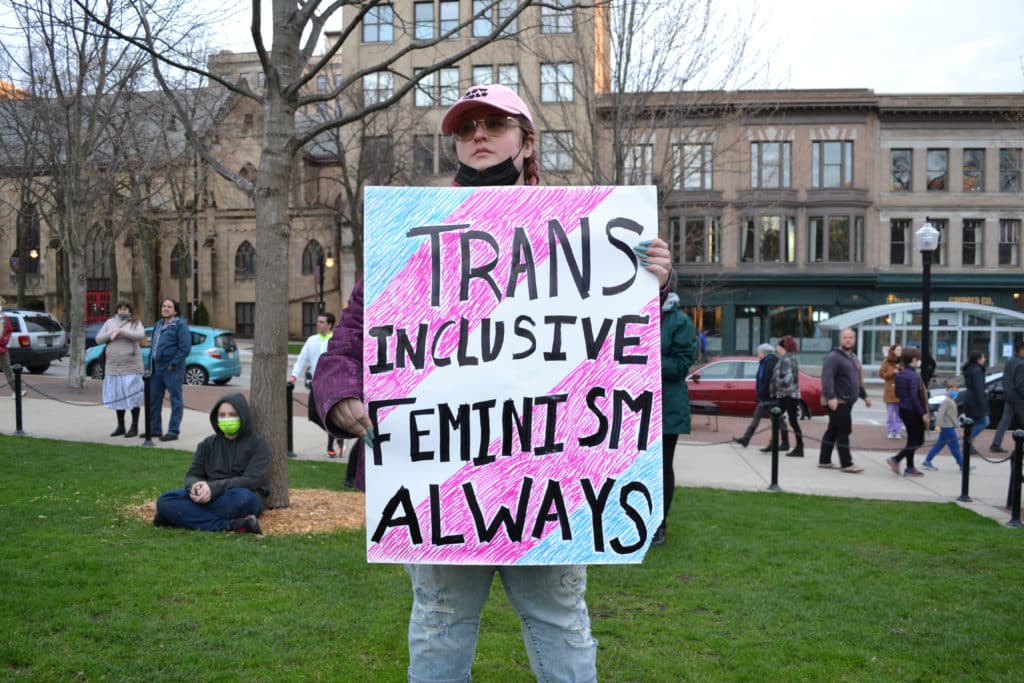 A person wearing a pink baseball cap stands facing the Capitol holding sign that reads "TRANS INCLUSIVE FEMINISM ALWAYS" with the pink and blue stripes of the Transgender Pride Flag drawn in the background. Photo by Hayley Sperling.