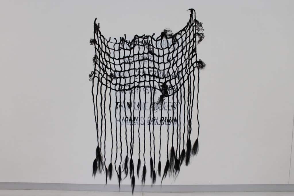 Teena Wilder’s "Again; The thin lines between witness and actor," a sculpture made of lengths of braided hair. It hands against a white wall on which a quote from James Baldwin, reading, "The line which separates a witness from an actor is a very fine line indeed," is rendered in wavy block letters.