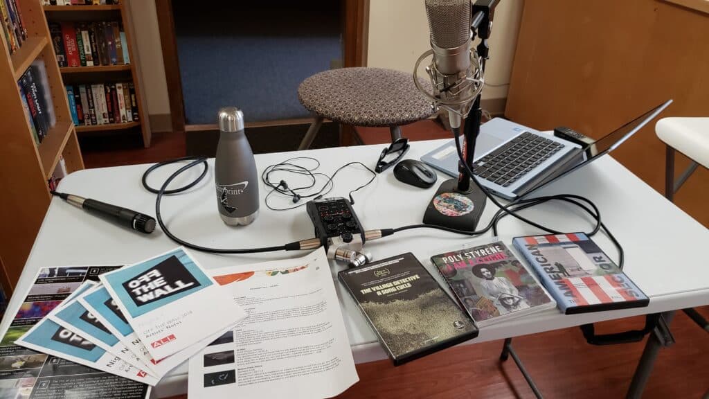 A photo of the summer podcast table setup at Four Star Video Rental. A Zoom Recorder sits on the center of the table, surrounded by microphones, a Chromebook, physical media of three Rooftop Cinema selections this summer, as well as programs from 2017 and 2018 screenings of "Off The Wall" at Arts + Literature Laboratory's old location.