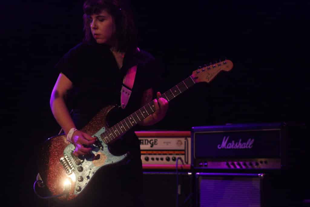 Scrunchies' guitarist/vocalist is shown in front of a Marshall stack and an Orange stack, playing a glittery Stratocaster. Her eyes are closed and she's mid-strum, to the left of the image. A light reflects brightly off of her guitar's input jack.