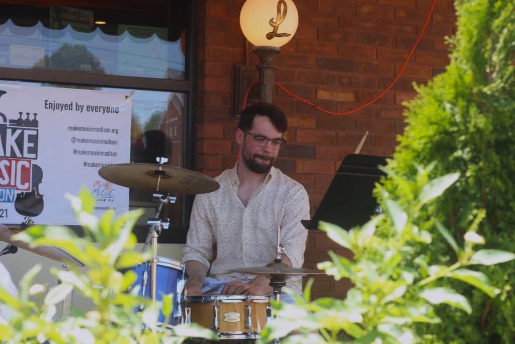 Lijario's drummer is pictured in front of Leopold's for Make Music Madison, smiling with his lips curled in. In the foreground, plants are shown in bokeh, branching out and providing an implicit frame as they cover the image's lower corners.