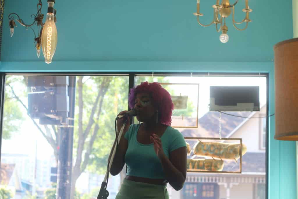 Vocalist Danielle Crim is shown singing in Mother Fool's during Make Music Madison, a view of the neighborhood is visible through the window that serves as her backdrop.
