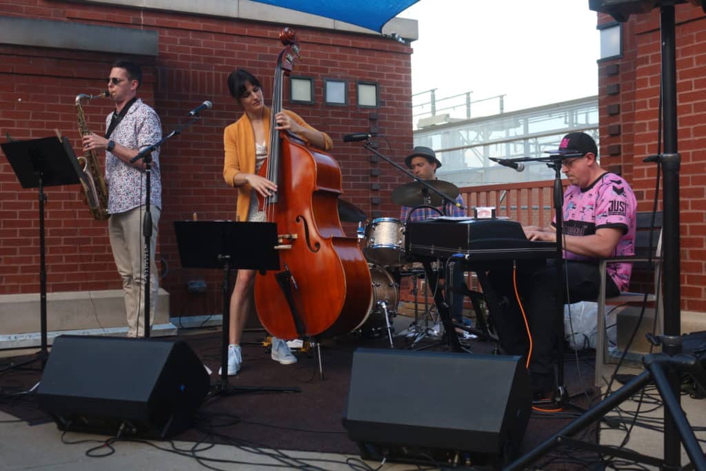 Inside Pocket is shown playing the High Noon Saloon's patio for Strollin' Capitol East, the kickoff event of Madison Jazz Festival. From left to right, there is a saxophonist, bassist, drummer, and keyboardist. The band's surrounded by brick buildings and playing underneath a blue overhang that cuts into the top middle of the shot.