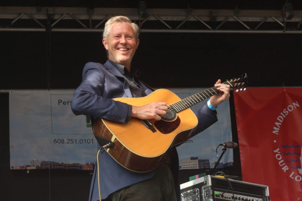 Robbie Fulks is shown standing straight up, holding his acoustic guitar, and grinning out at the crowd during Marquette Waterfront Festival.