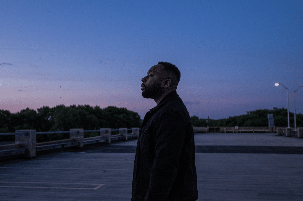 A Black man stands near the edge of a body of water. His silhouette is dark and the image's palette is awash in dark blues and a streak of orange and purple pastels above the tree line on the far end of the water. The man's looking straight ahead to the image's left, creating a perfect profile. 