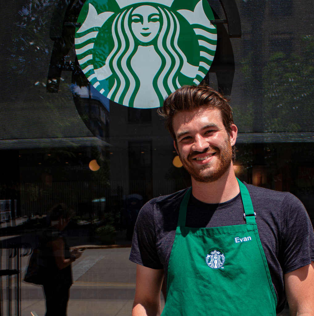 A Starbucks worker is shown standing outside the chain's Capitol Square location, wearing a green apron and smiling toward the camera.