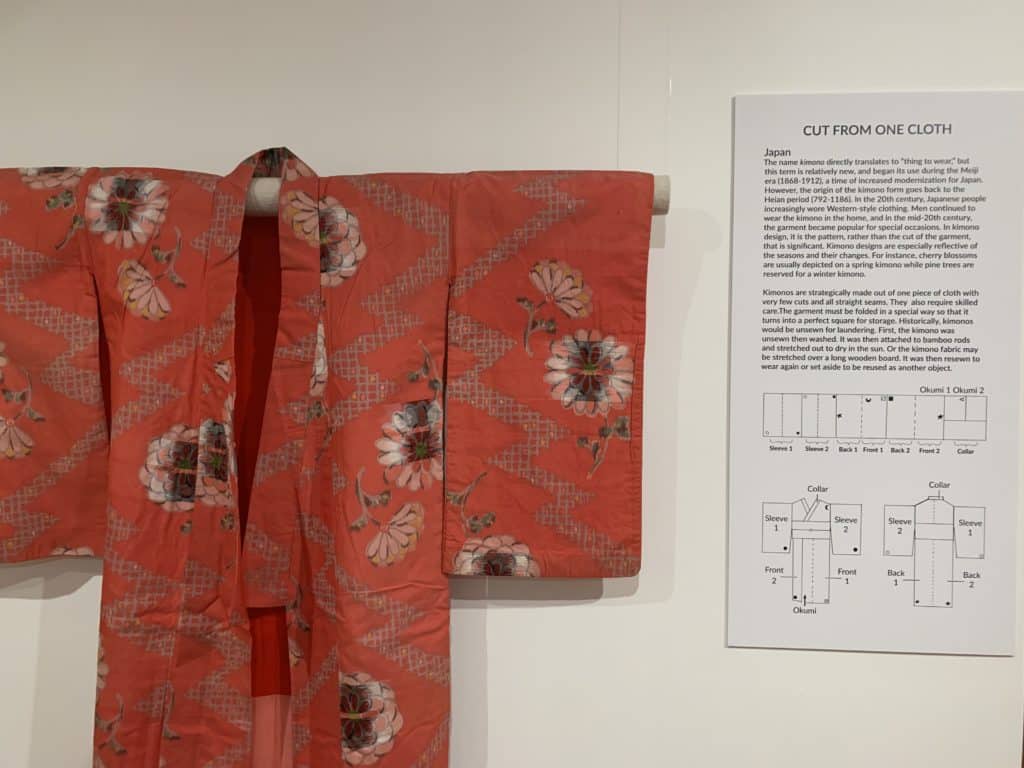 A kimono is shown hanging on a wall in a gallery. Next to it is a gallery sign with the headline "Cut from one cloth," which contains passages of smaller text and a diagram showing how the kimono is made.