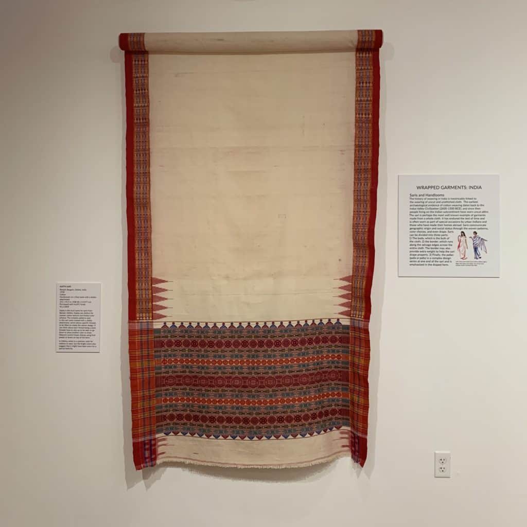 A photo shows a Sari hanging on a wall in a gallery. Next to it is a small gallery sign with the headline "Wrapped Garments: India."