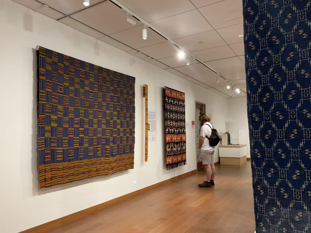 A photo shows a large piece of Kente cloth hanging on a wall in a gallery. In the background is a gallery visitor looking at another piece.