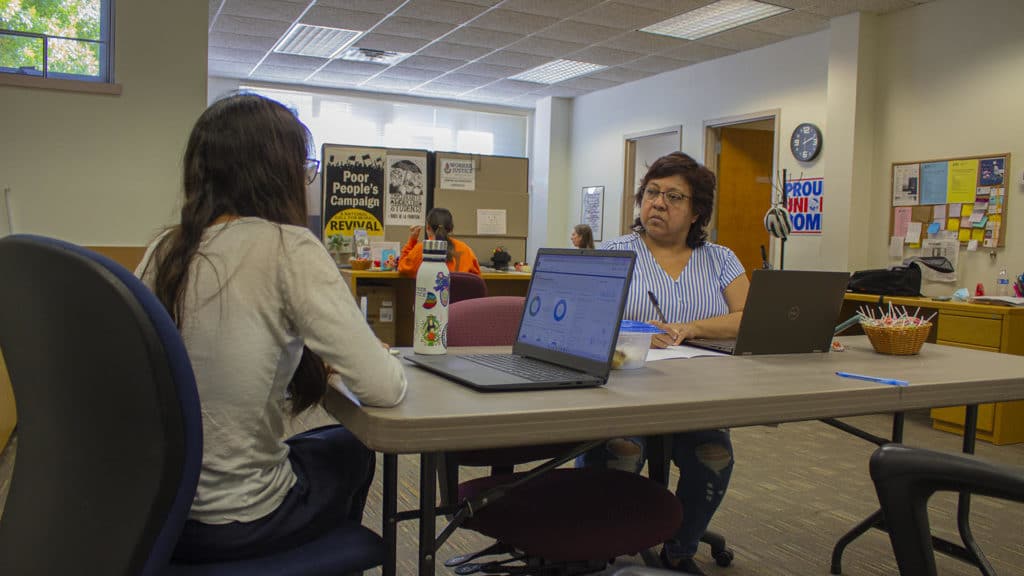 Frida Ballard (left) and Socorro Cortez (right), shown sitting at a table in an office with colorful posters and postings in the background, discuss their work as organizers for Worker Justice. The two converse as much in Spanish as English. Many of the staff are bilingual.