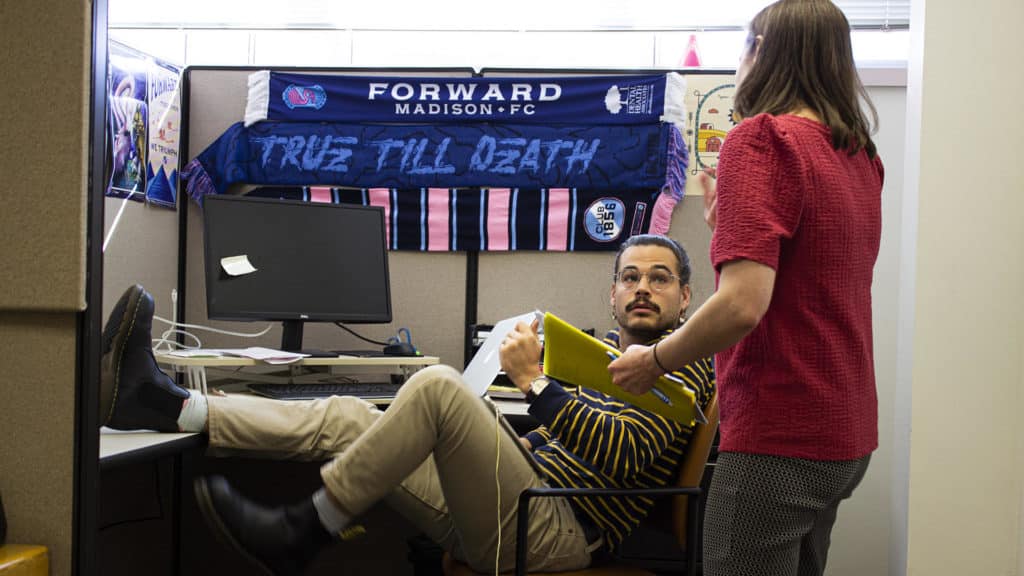Program Director Robert Christl and Faith Outreach Coordinator Gretchen Baumgardt discuss upcoming dealings with faith communities in Christl’s cubicle which drips with the iconography of the local soccer club, Forward Madison FC. Christl is shown sitting in an office chair with one foot propped up on a desk, while Baumgardt stands in the foreground talking with him and gesturing.
