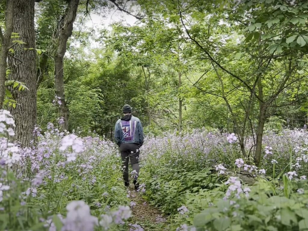 A still from the "Grown Used To It" video in which Tyler Fassnacht is pictured walking away from the camera in lush green woodlands. Flowers grow all around him as trees tower above.