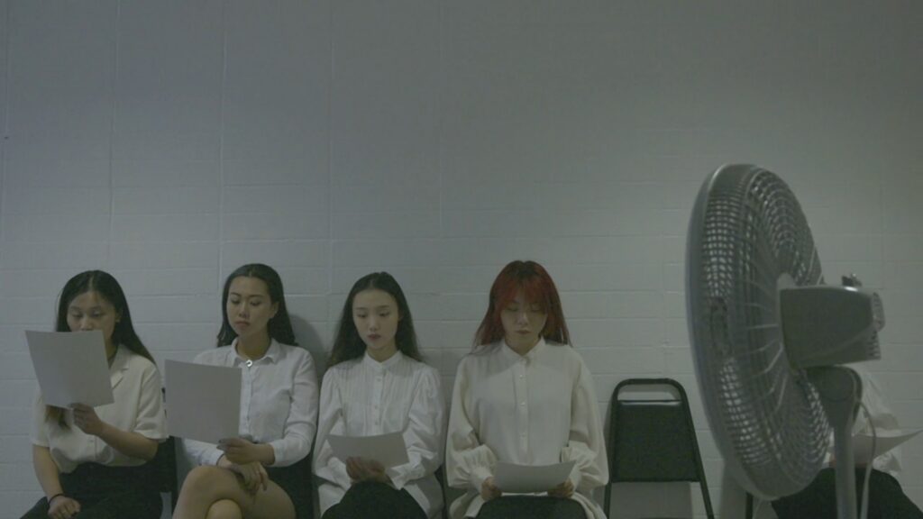 In the second shot of the film, four similarly dressed Asian women sit outside a casting room in a blank hallway, studying the script with a blank expression, and waiting their turn to audition.
