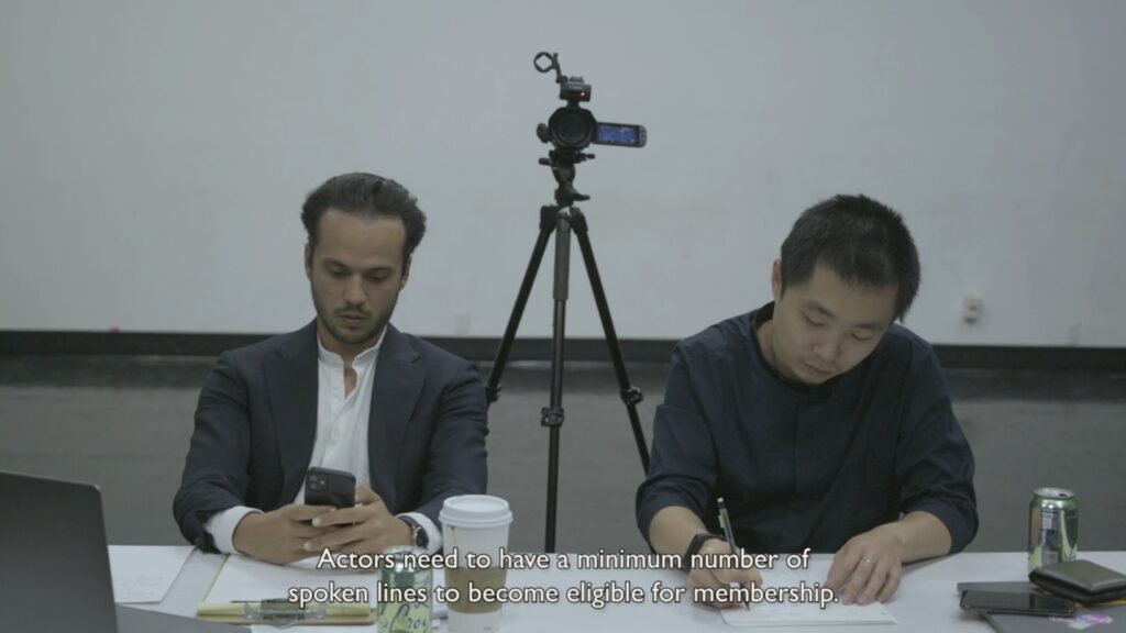 A disinterested casting director (Paul Sohm, left) checks his smart phone after fielding a subtitled question from an actress about whether the action in the script officially counts as a line. To the right, his translator (Xuege Cai) makes a note.