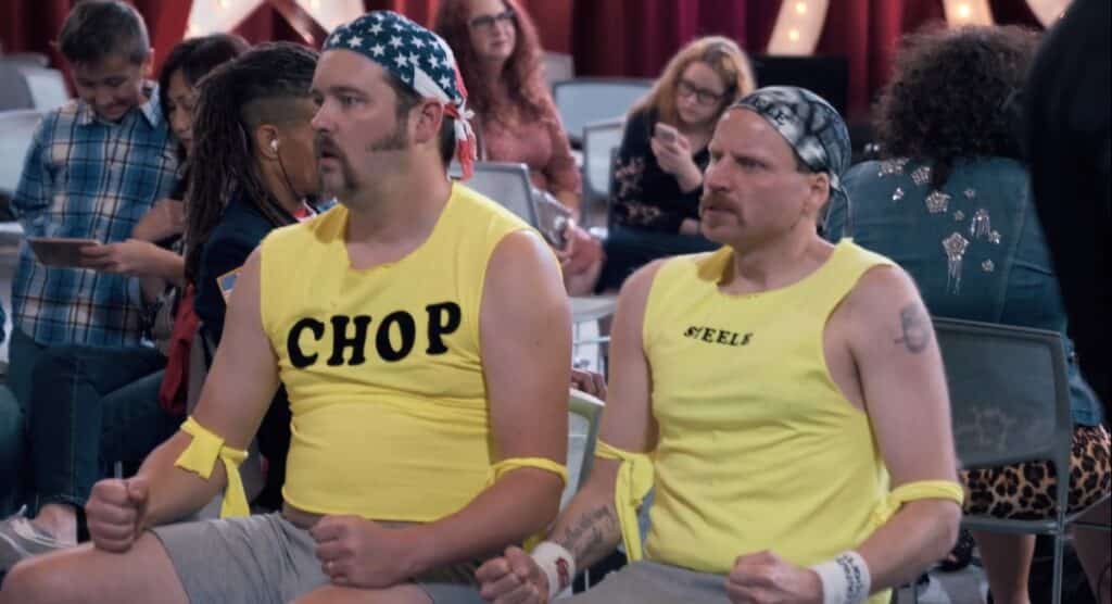 Joe Pickett and Nick Prueher sit in a waiting area while dressed as their strongman characters Chop and Steele. Pickett looks exhausted while Prueher looks angry and determined.