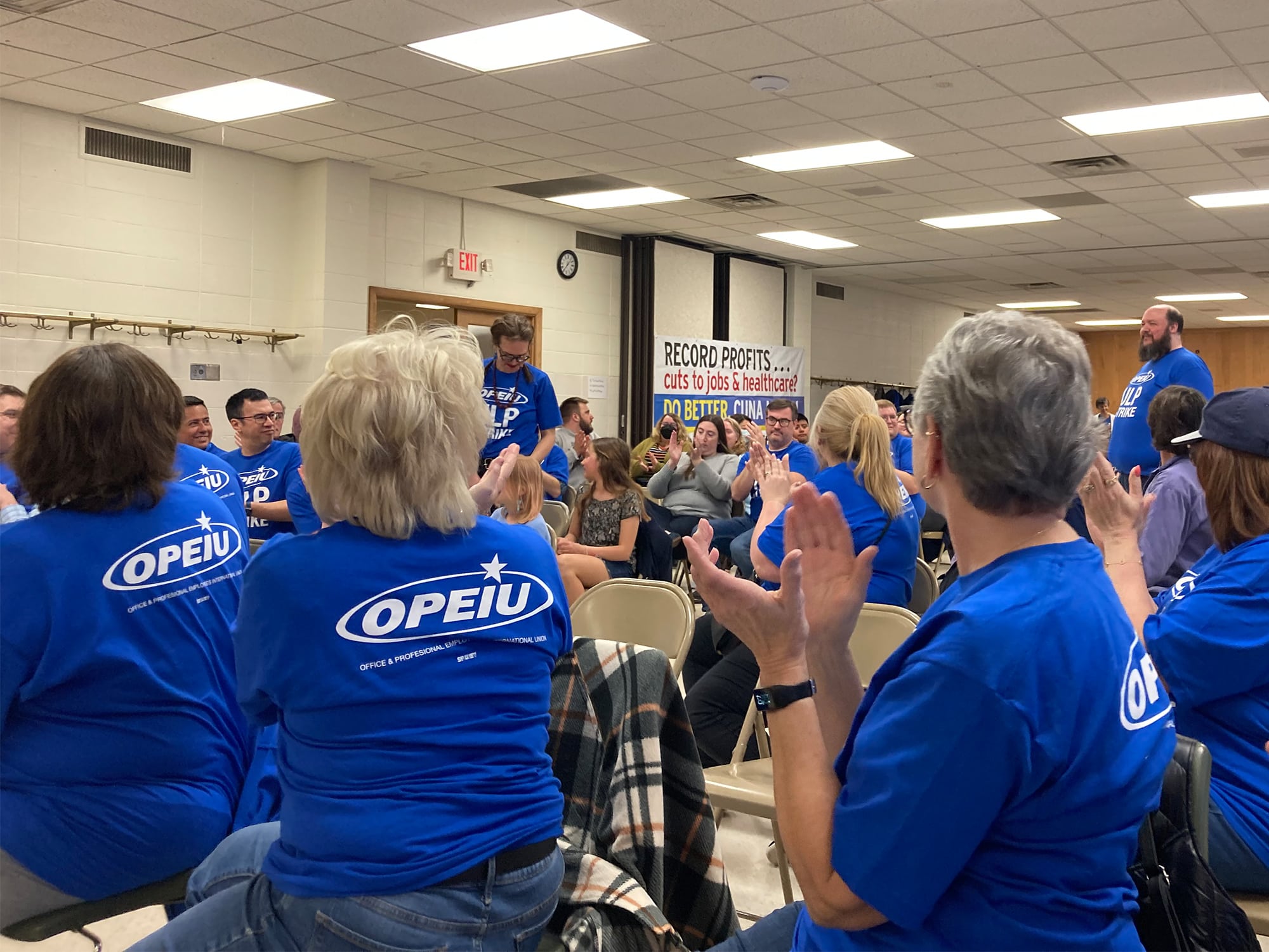 A photo shows a group of workers, all wearing blue shirts with the Office and Professional Employees International union logo on the backs, applauding in a meeting hall. A sign visible near the back of the room reads, “Record profits… cuts to jobs and healthcare? Do better, CUNA Mutual.”