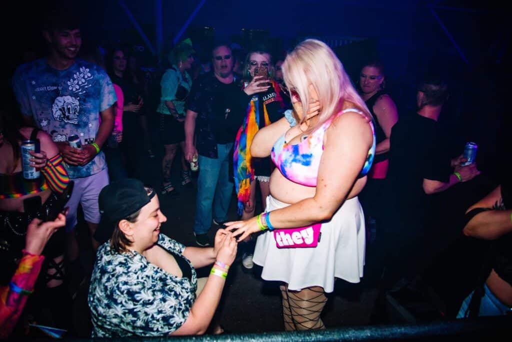 This photograph shows Olivia Steele on one knee on a club dance floor putting an engagement ring on her partner Charli's finger as Charli covers her mouth with her other hand out of emotion.