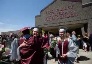 Graduating students in caps and gowns hug in front of the entrance to Fox Valley Lutheran High School.