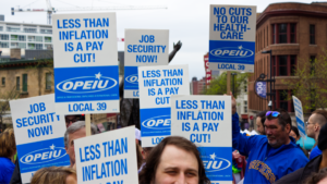 A photo shows an assortment of OPEIU Local 39 signs with slogans such as "Less than inflation is a pay cut" and "Job security now."