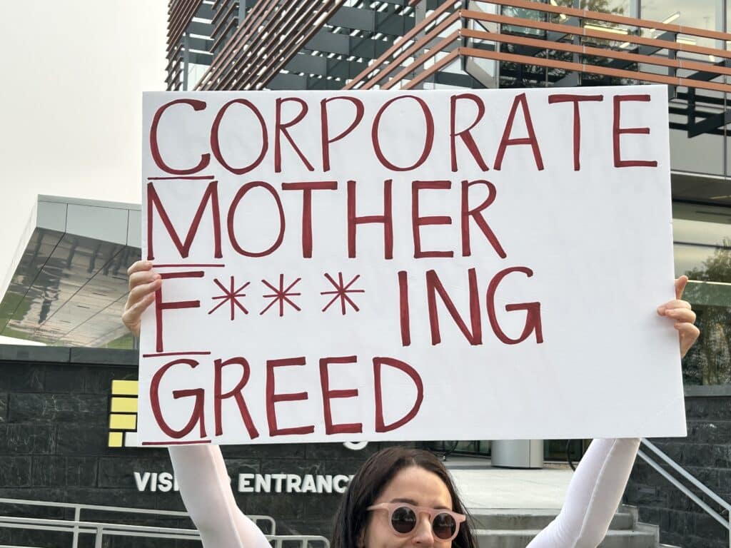 A photo shows a picketer holding a hand-lettered sign reading "CORPORATE MOTHER F***ING GREED."