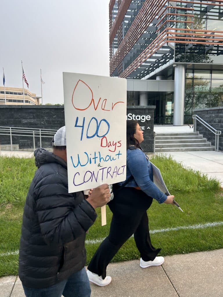 A photo shows a picketer carrying a hand-lettered sign that reads "Over 400 days without a contract."