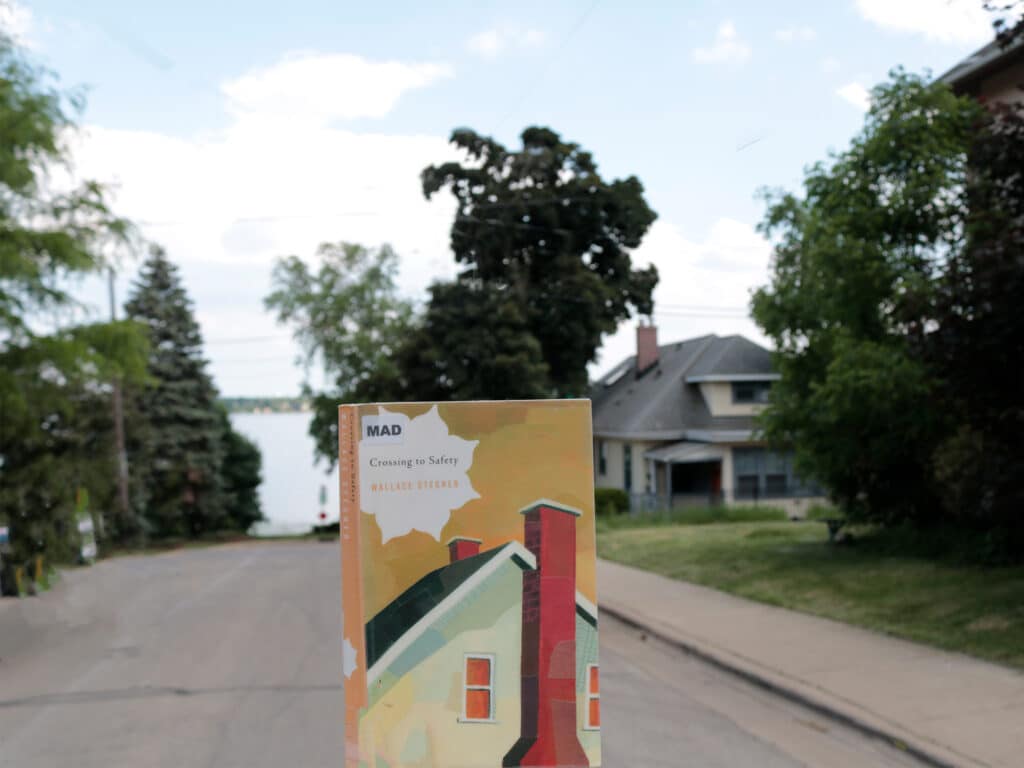 A photo illustration shows a paperback copy of Wallace Stegner’s “Crossing To Safety” held up in the foreground, with a residential Madison street and the shore of Lake Mendota visible in the background.