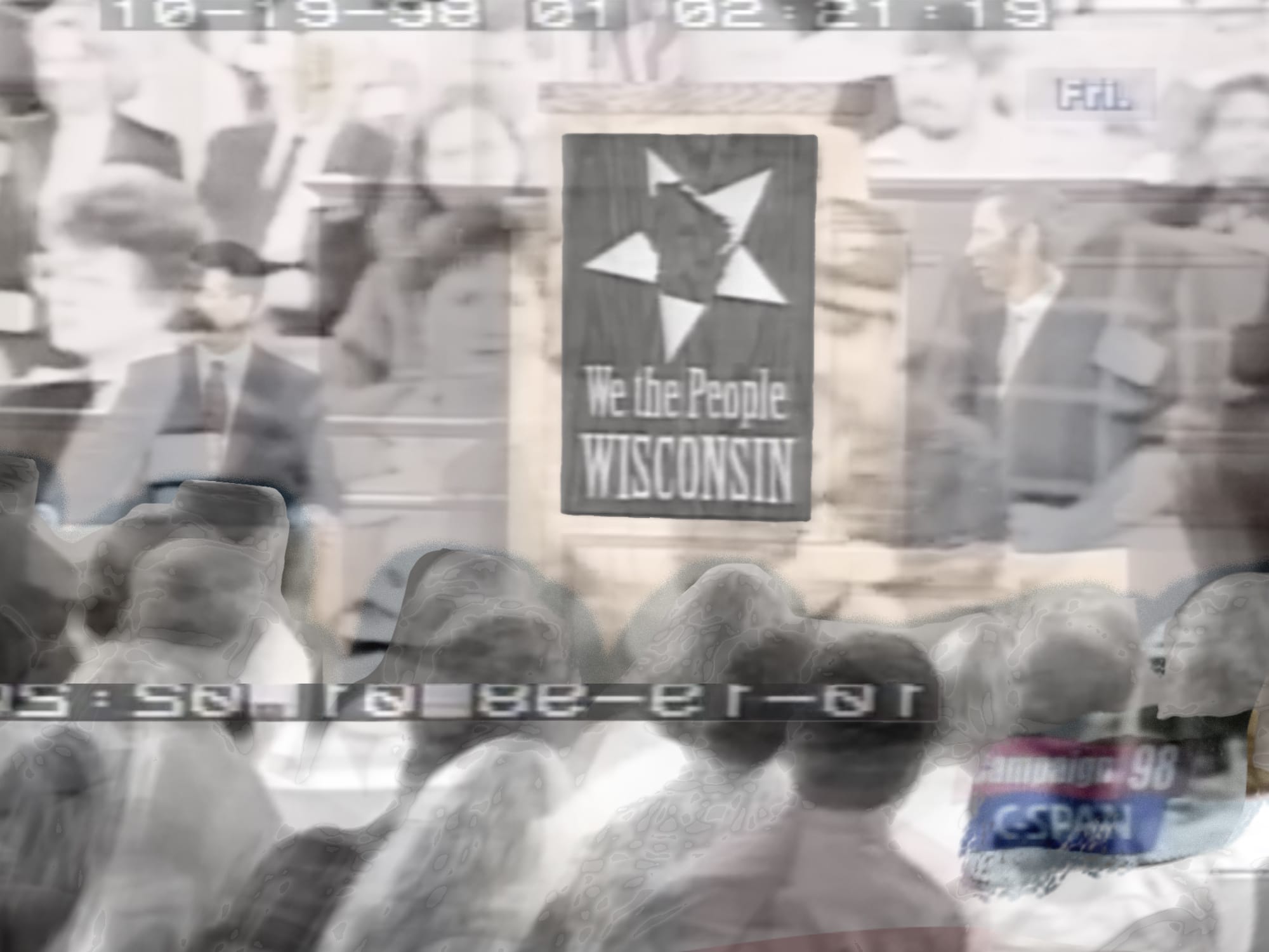 A collage layers screenshots from a 1998 town-hall debate between U.S. Senate candidates Russ Feingold and Mark Neuman. A view of a seated crowd from behind is visible, as is a faint outline of another perspective that faces the crowd from the front of the room, and the two candidates standing behind podiums. Between the two candidates is a large sign that reads “We the People Wisconsin.” Also visible are video time stamps. All the elements are filtered through distortion and black-and-white coloration.
