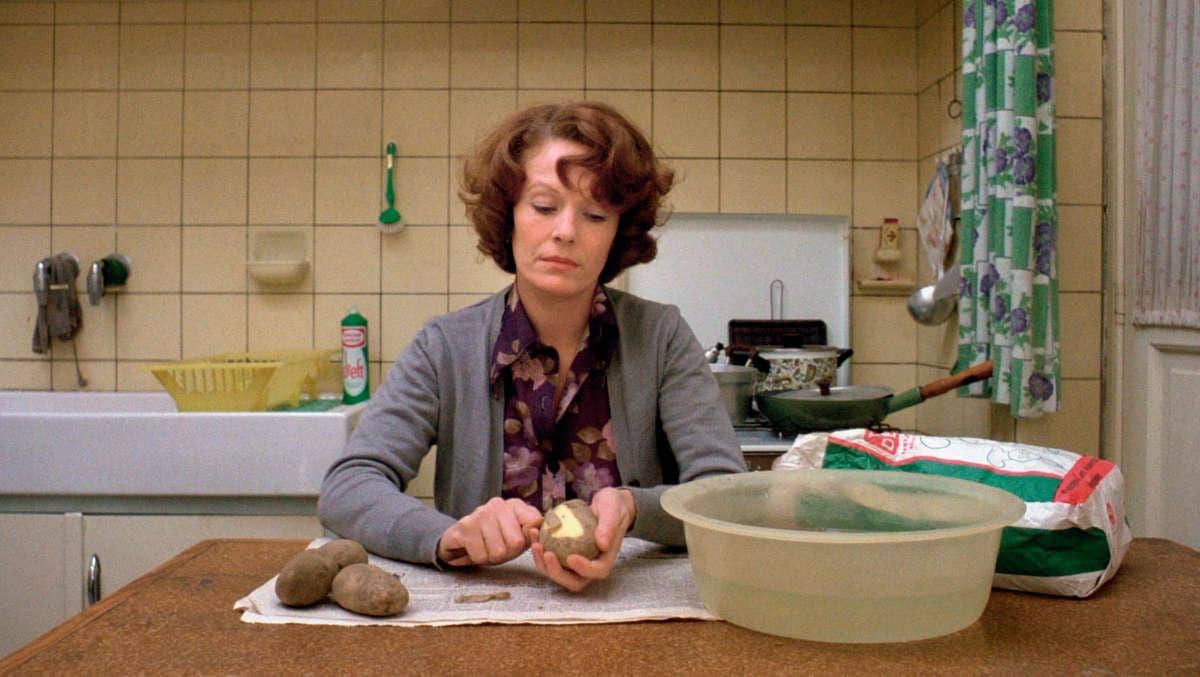Jeanne Dielman peels potatoes at a kitchen table with a neutral expression.