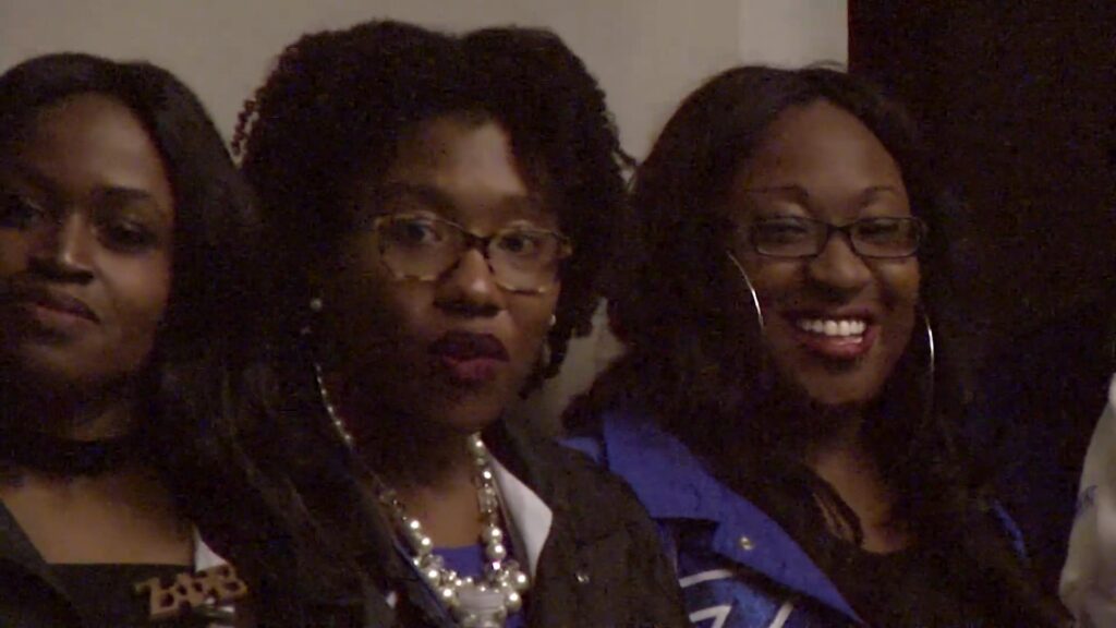 Three Black women turn their gaze towards a camera filming an event at Tau Theta Chapter of Zeta Phi Beta.