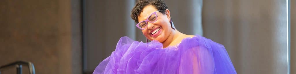 Dr. Sami Schalk smiles at the camera while wearing lilac cat-eye glasses, silver triangle earrings, and a puffy tulle purple dress.