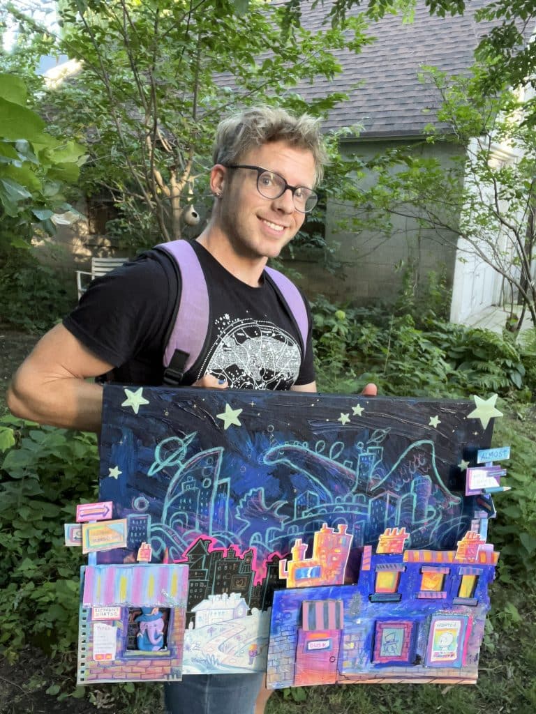 A photo shows artist Will Santino holding up the illustrated mixed-media work pictured at the top of the story. He is shown standing in front of some bushes and facing the camera.