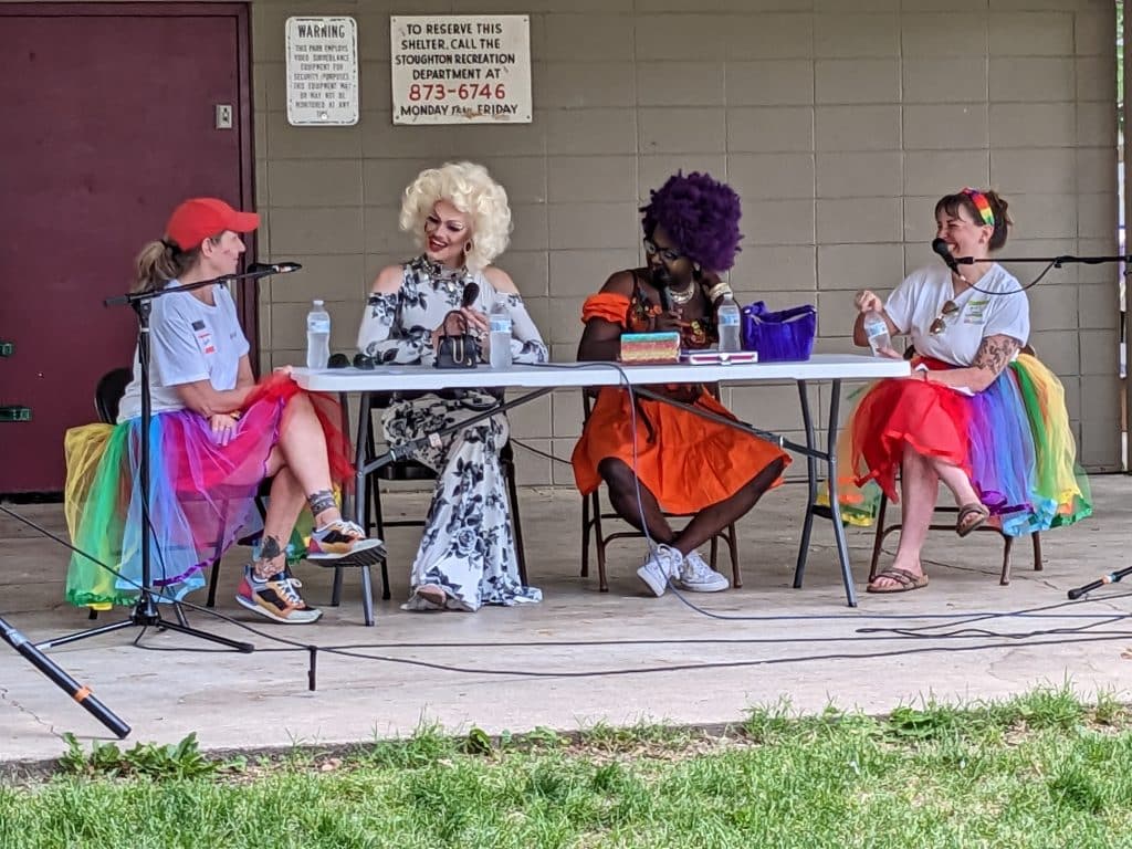 Photo of an event at an outdoor shelter where two librarians in rainbow tutus sit at a long table with two drag queens.