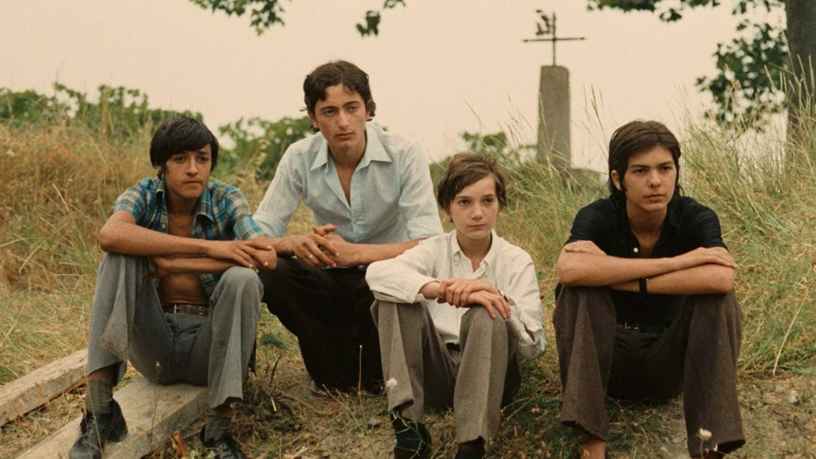 Four teenage boys sit on a sparsely grassy hill, all staring intently to the right of the frame. Daniel, the youngest and smallest among them, wears a white button shirt with long sleeves.