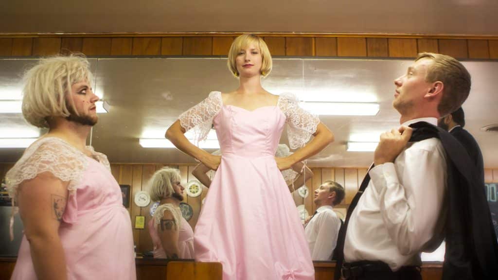 Cynthia Burnson is centered in the image, her hands balled into fists that are resting on her hips. She's standing up straight and looking directly at the camera. Shane O'Neill is to the left of the image, looking up at Burnson. To Burnson's right are the band's rhythm section. The picture's composed symmetrically, and the band's posing in front of a mirror. Their reflections fill the gaps between the members. Burnson and O'Neill are wearing matching pink dresses with white lace sleeves, while the other band members are wearing traditional suit-and-tie ensembles.  