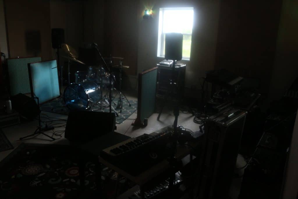 A shot of Mobius Glen's live room shows drums with transparent blue shells on top of a rug near the back right corner of the room. Near the bottom of the image is a row of synths and equipment cases. Various speakers can be seen either mounted or on the ground. Audio panels can be seen meticulously positioned around the drums. 