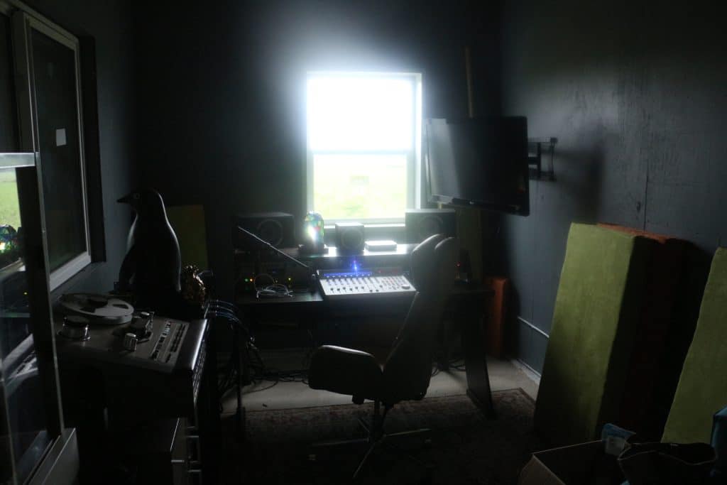 A wide view of Mobius Glen's control room shows an assortment of recording equipment, a small penguin statue perched on a desk in the left of the image, and a window near the back wall. All of the playback devices are near the window. 