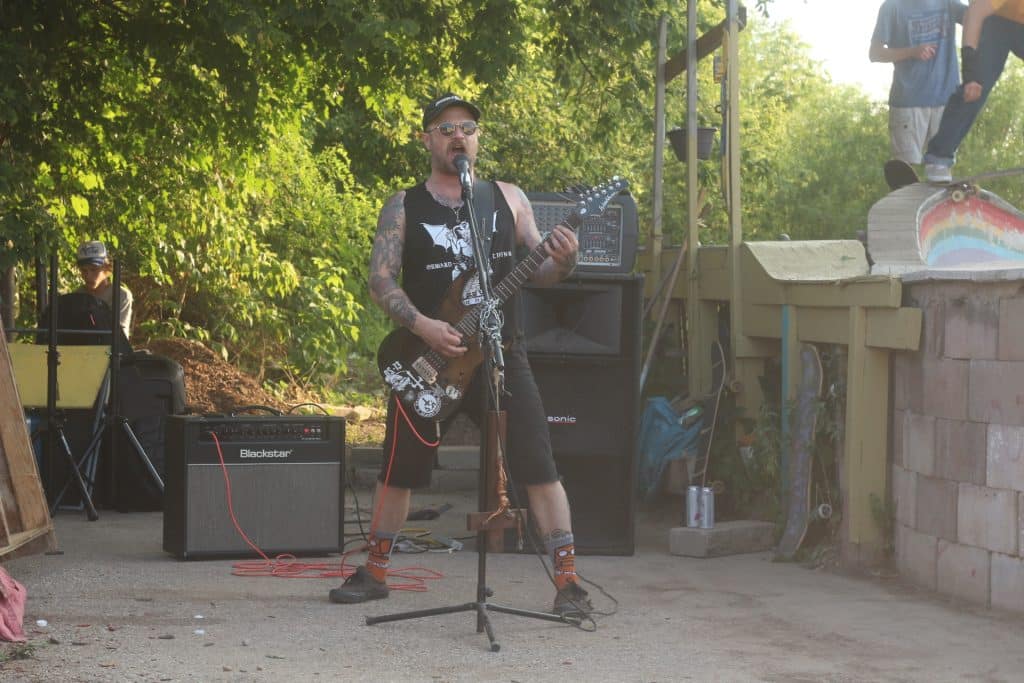 Infernal Trench plays at DIY skatepark Forward Living. He's wearing sunglasses, a black tank top, black jean shorts, Toy Machine socks, and black shoes. He's playing a black guitar and singing into a microphone. In the upper right corner of the image, someone can be seen dropping in on their skateboard. In the background to his left, there is a person running sound. 