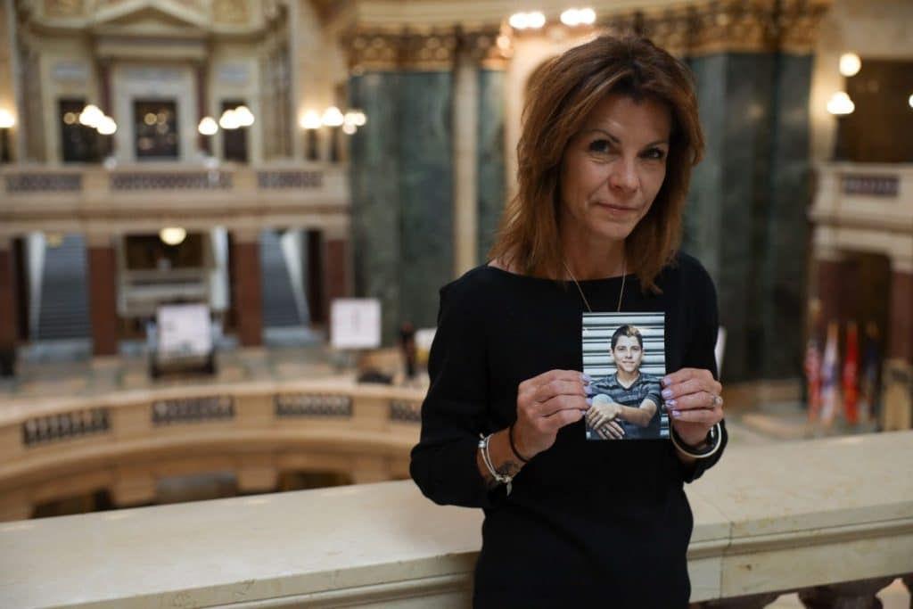 Photo of Sheila Lockwood holding a photo of a young man, the rotunda of the state Capitol behind her. 
