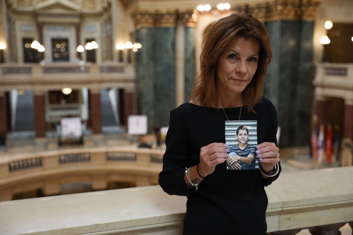 Photo of Sheila Lockwood holding a photo of a young man, the rotunda of the state Capitol behind her.