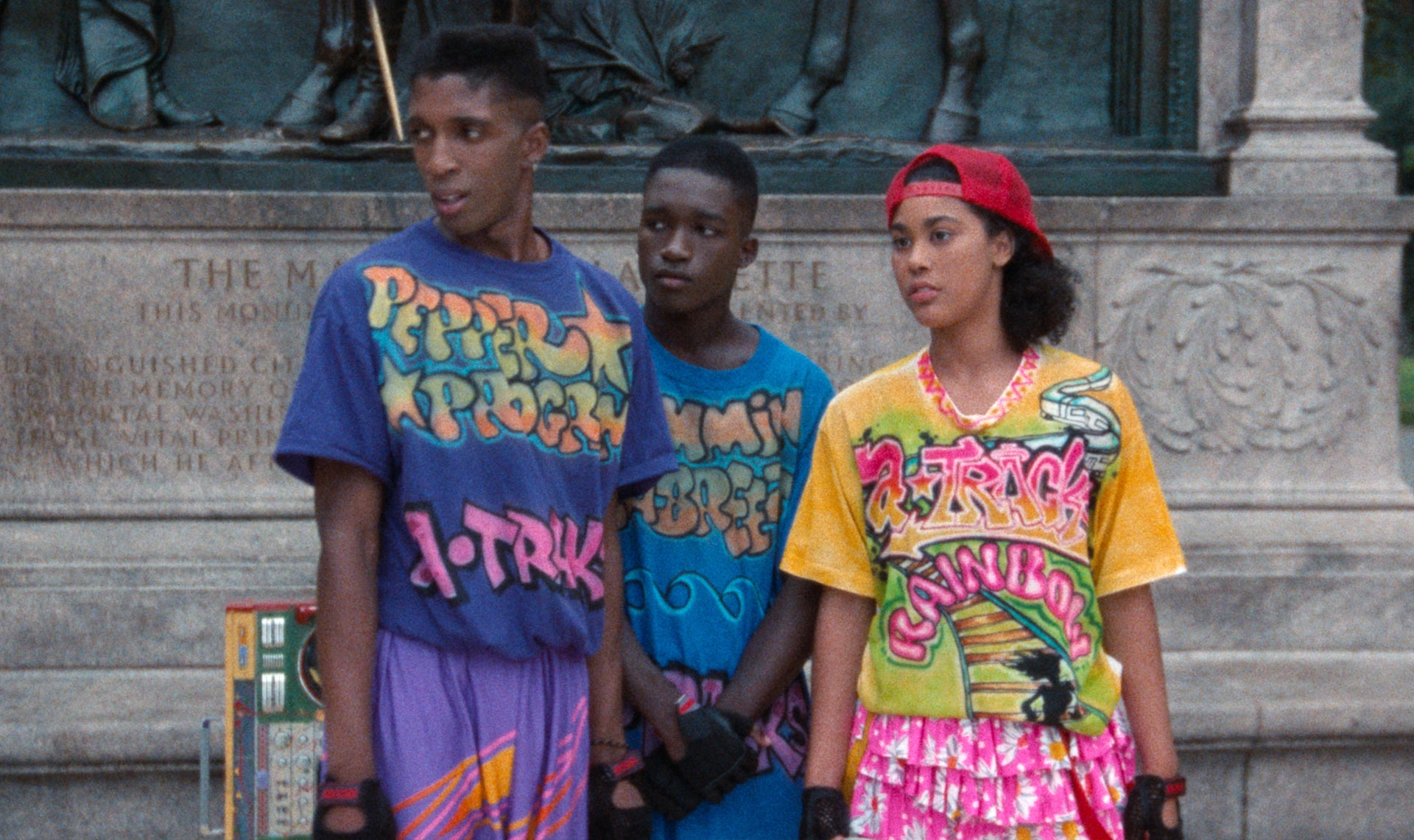 Three Black teenagers in colorful, custom-made clothing that displays their names and "A-Tracks" stare off to the left in front of the Lafayette Memorial in Prospect Park Brooklyn. Their boombox sits on the ground behind Pepper, the tallest of the A-Tracks team. Rainbow stands out on the right in her yellow and pink attire and red backwards hat.