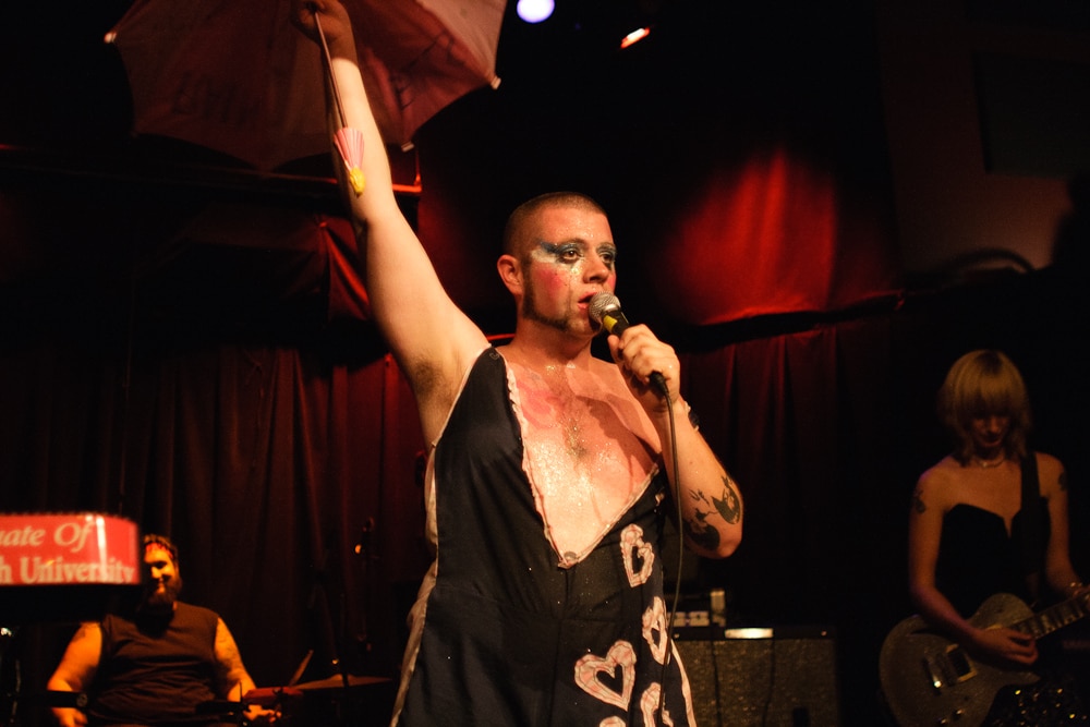 Screamin' Cyn Cyn And The Pons are performing live. Vocalist Shane O'Neill is centered in the image, wearing a black dress with a plummeting neckline. O'Neill's wearing heavy eye makeup and raising his left hand to the sky, while using his right hand to clutch the microphone while singing. Cynthia Burnson's playing guitar on the right side of the image, while The Pons Pons' drummer is smiling in the lower left corner. 