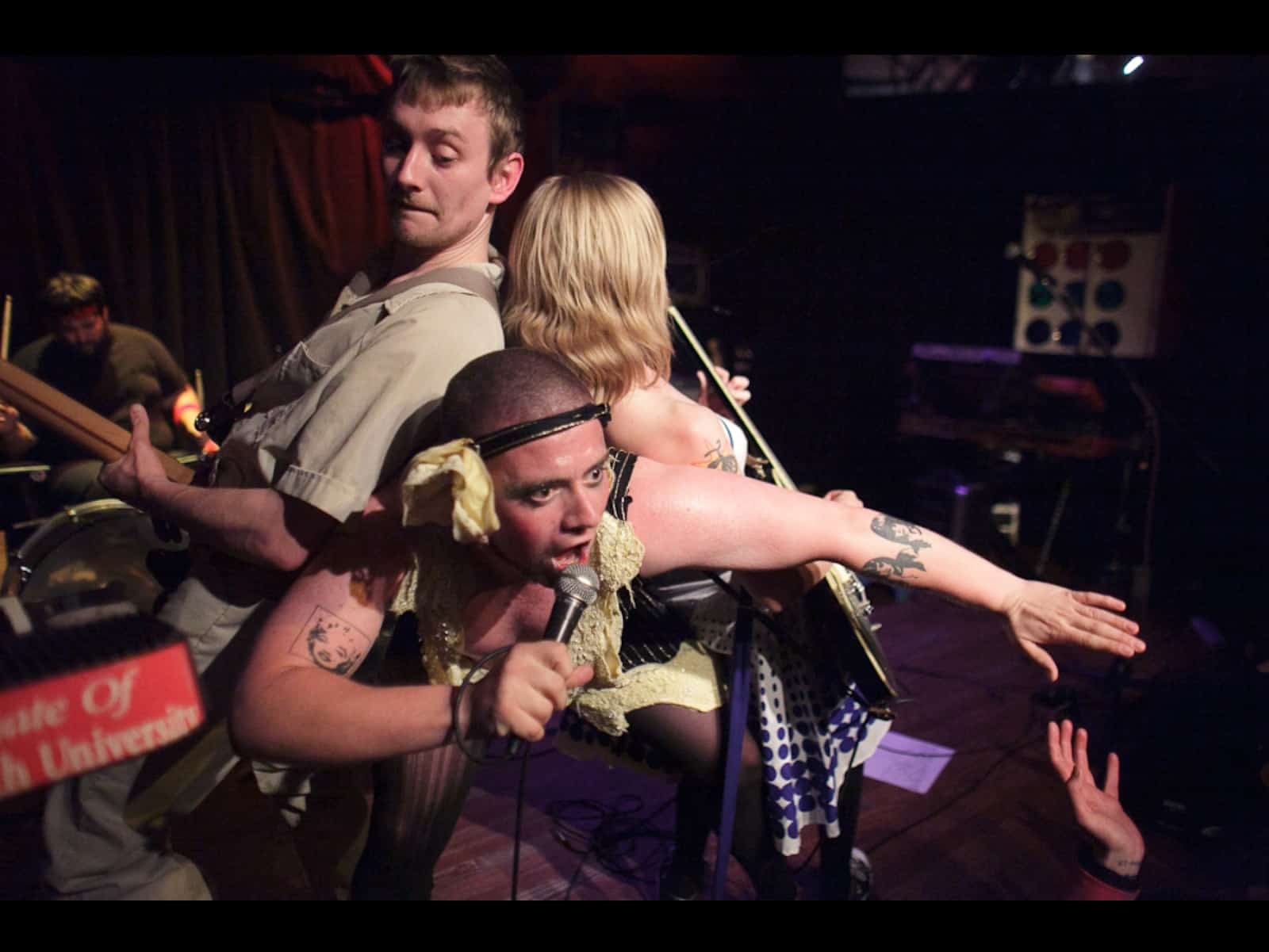 The members of Screamin' Cyn Cyn And The Pons are shown mid-performance. The backdrop is black. Vocalist Shane O'Neill is in the bottom of the image, wide-eyed, and wearing a makeshift headband. O'Neill's free arm is extended out to the lower right corner of the image. An audience member's hand is reaching out towards his arm. Leaning on O'Neill are Cynthia Burnson and The Pons Pons' bassist. Visible towards the back of the stage and tucked into the upper left corner of the image is The Pons Pons' drummer.