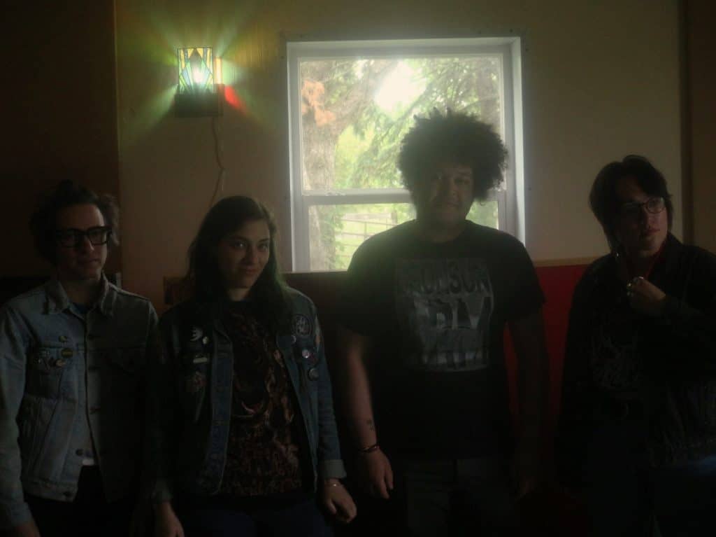 The members of Cult Of Lip are shown in the low light of Mobius Glen, their studio and practice space. They are standing against a wall, just underneath a window. A stained glass lamp is affixed to the wall in the upper left hand corner of the image. From Left to Right: Ronnie Lee, Hannah Porter, Terrance Barrett, and Emili Earhart. 