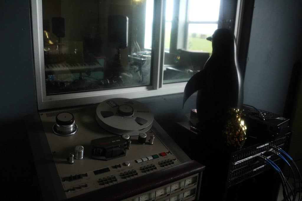 Recording equipment sits at the bottom right of the image, to its left is a small statue of a penguin. The penguin's positioned so that it is looking through a window from its place in a studio control room to the live room on the other side of the wall. 
