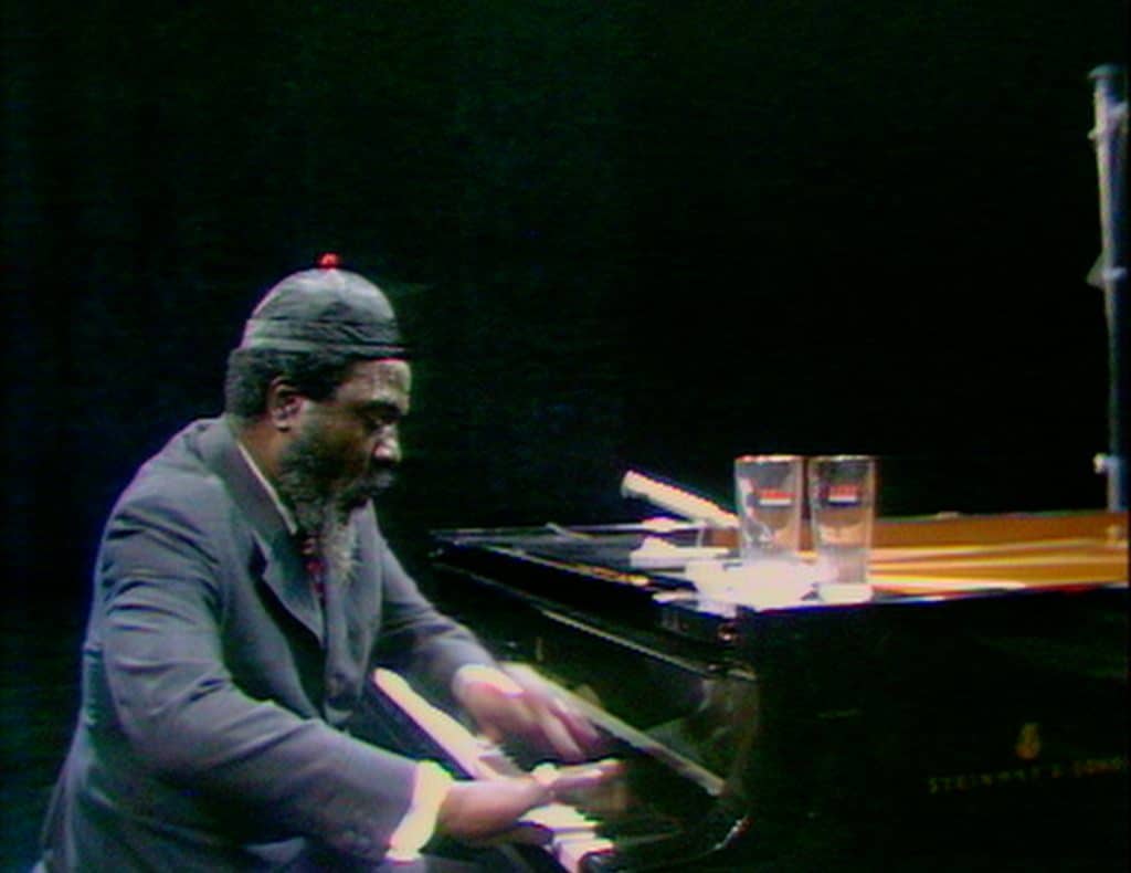 Against a TV program's black background, jazz pianist Thelonious Monk sits at a piano that has two glasses of water on the edge of the lid. The image captures him playing intensely, mid-motion.